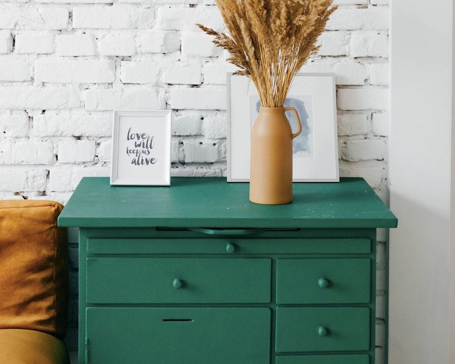 A green chest of drawers with dark yellow vase and white picture frames on top standing in front of a white wall.