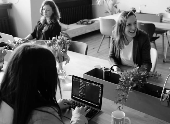 Three women sitting at a table in an office setting. Two are working on their computers, one is looking left and smiling